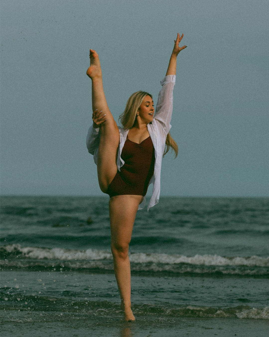 Woman performing a high leg extension on the beach, wearing a maroon swimsuit and white shirt, with waves in the background.